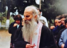 an old man with a long white beard wearing a cross on his arm and standing in front of other people