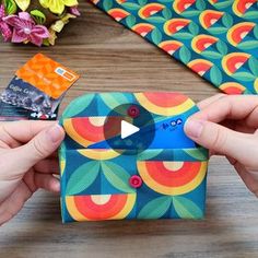 two hands holding an origami box with a video game on it and some flowers in the background