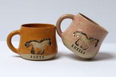 two mugs with horses painted on them sitting next to each other, one is brown and the other is tan