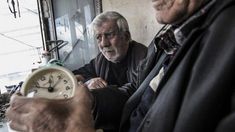 two older men sitting next to each other in front of a wall with an old clock on it