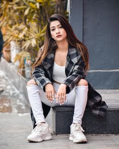 a woman is sitting on a bench wearing white pants and a plaid coat, with her hands in her pockets