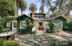 a green house surrounded by trees and greenery in the front yard with a picnic table on the patio