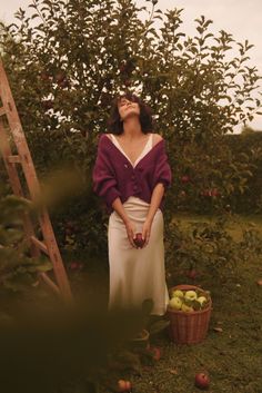 a woman standing in front of an apple tree with her eyes closed and apples in the basket