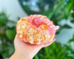 a hand holding a small crocheted donut in front of a green plant