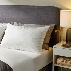 a bed with white sheets and pillows next to a night stand, lamp and nightstand