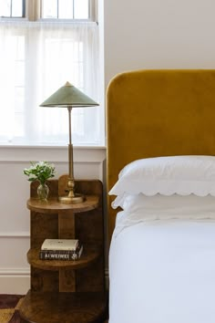 a bed with a yellow headboard next to a lamp on a night stand in front of a window