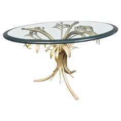 a round glass table with gold leaves on it's base and an oval glass top