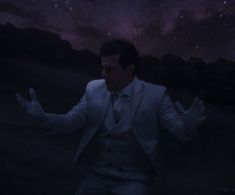 a man in a white suit and tie with his hands out to the stars above him