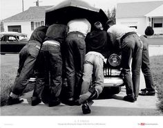 four men are huddled under an umbrella on the back of a car in front of a house