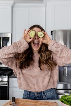 a woman holding two slices of avocado in front of her eyes while standing next to a cutting board