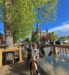 a woman sitting at a table on a deck in front of a tree and buildings