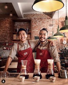 two men in aprons standing next to each other at a counter with coffee cups