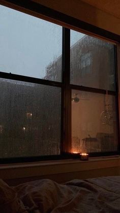 a bedroom with a window that has rain coming down the outside and candles lit up in front of it