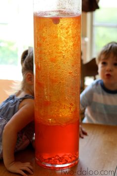 two young children sitting at a table with a tall glass vase filled with orange liquid