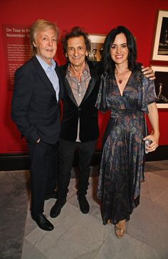 two men and a woman standing next to each other in front of a red wall