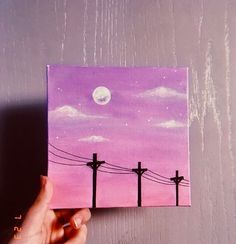 a person holding up a small painting with power lines and the moon in the background