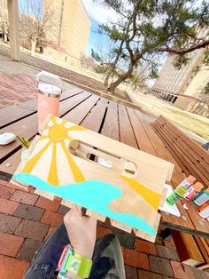 a person sitting at a picnic table holding up a piece of wood with paint on it