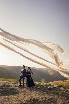 a man and woman standing on top of a hill next to a white scarf flying in the air