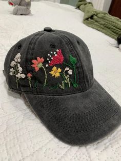 a gray hat with embroidered flowers on it sitting on a bed next to a stuffed animal