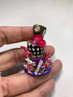 a hand is holding a miniature hat with flowers and other things on it's top