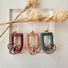 three different colored earrings hanging from hooks on a white surface next to dried flowers and stalks