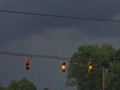 two traffic lights hanging from wires in front of trees on a cloudy day with dark clouds overhead