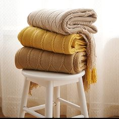 three blankets stacked on top of each other in front of a white stool and window