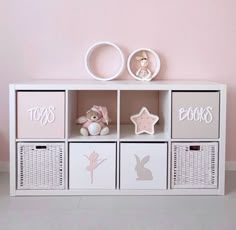 a white shelf with four bins and some toys on it in front of a pink wall