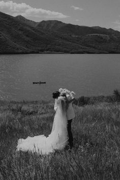 a bride and groom are standing in front of a body of water with mountains in the background