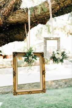 an old frame hanging from a tree with flowers and greenery on the front hung by ribbons