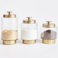 three glass jars with gold lids are sitting side by side on a white surface, one containing flour and the other containing powder