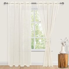 a window with white curtains and a vase next to it on a wooden table in front of the window