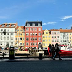 people walking on the sidewalk in front of some colorful buildings with boats docked at the dock