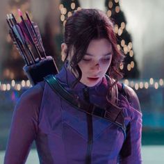 a woman holding an arrow in her right hand and looking down at the ground with christmas lights behind her