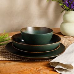 a stack of green plates sitting on top of a wooden table next to a vase with purple flowers