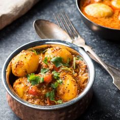 a bowl filled with potatoes and curry next to a silver spoon on top of a table