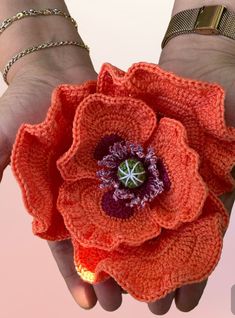 two hands holding an orange crocheted flower with chain bracelets on it's wrist