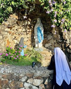 a statue of the virgin mary and child jesus in front of a stone wall with purple flowers