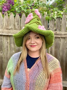 a woman wearing a knitted hat with flowers on it's brim, standing in front of a fence