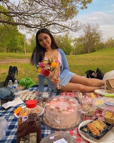 a woman sitting at a picnic table with cake and flowers in her hands, surrounded by other food items