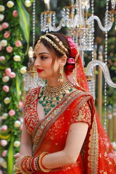 a woman in an orange and red bridal outfit with jewelry on her head, standing next to flowers