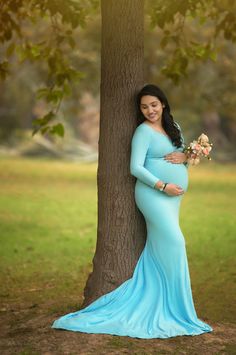 a pregnant woman leaning against a tree in a blue dress with her hands on the belly