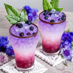 two glasses filled with blueberries and ice sitting on top of a table next to purple flowers