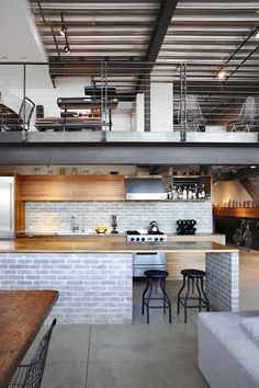 an open kitchen and living room area in a loft style home with exposed ceilings, white brick walls, stainless steel appliances and bar stools