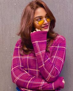 a woman wearing glasses and a pink sweater poses for a photo in front of a gray wall