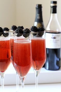 three glasses filled with liquid and blackberries next to two bottles of wine on a counter