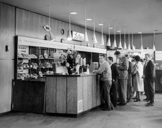 black and white photograph of people standing in line at a store counter with hats hanging from the ceiling