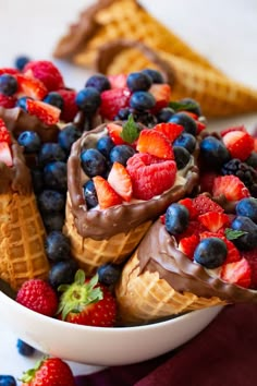 a bowl filled with ice cream covered in berries and chocolate sauce next to waffle cones