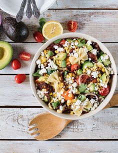 a white bowl filled with pasta salad next to sliced avocado and cherry tomatoes