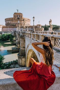 a woman in a red dress is sitting on the edge of a bridge looking at an old castle